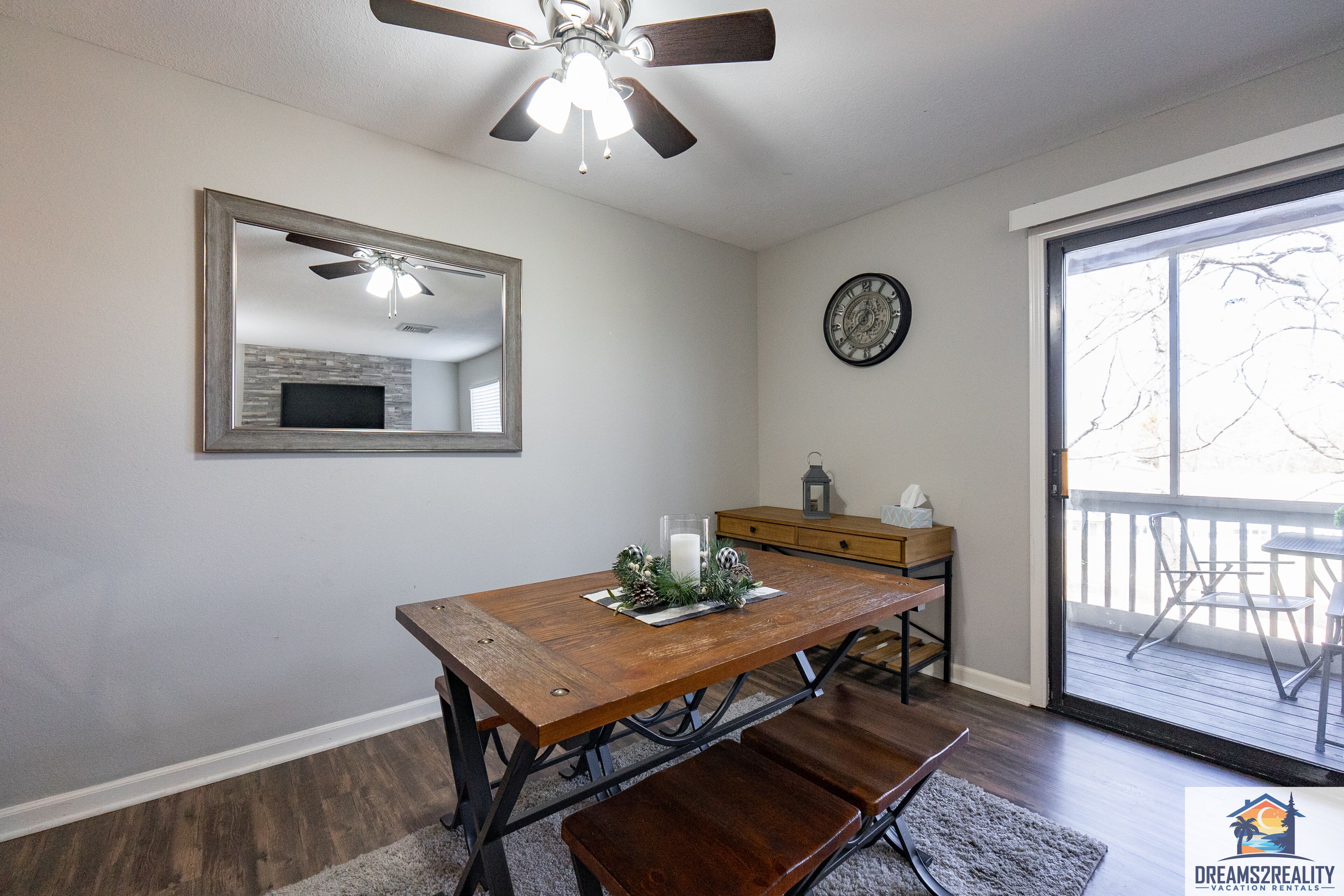 A dining area with a wooden table, benches, a ceiling fan, a fireplace, a clock, and a view of the outdoors through a sliding glass door.