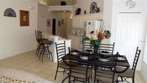 A dining area with a table set for four, surrounded by chairs. A kitchen is visible in the background with a refrigerator and various appliances. - Private Pool & Spa • Game Room • 2 King Ensuites • 15 Minutes to Disney (Clermont)