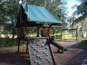 A wooden play structure with a slide and climbing area, surrounded by trees and gravel.