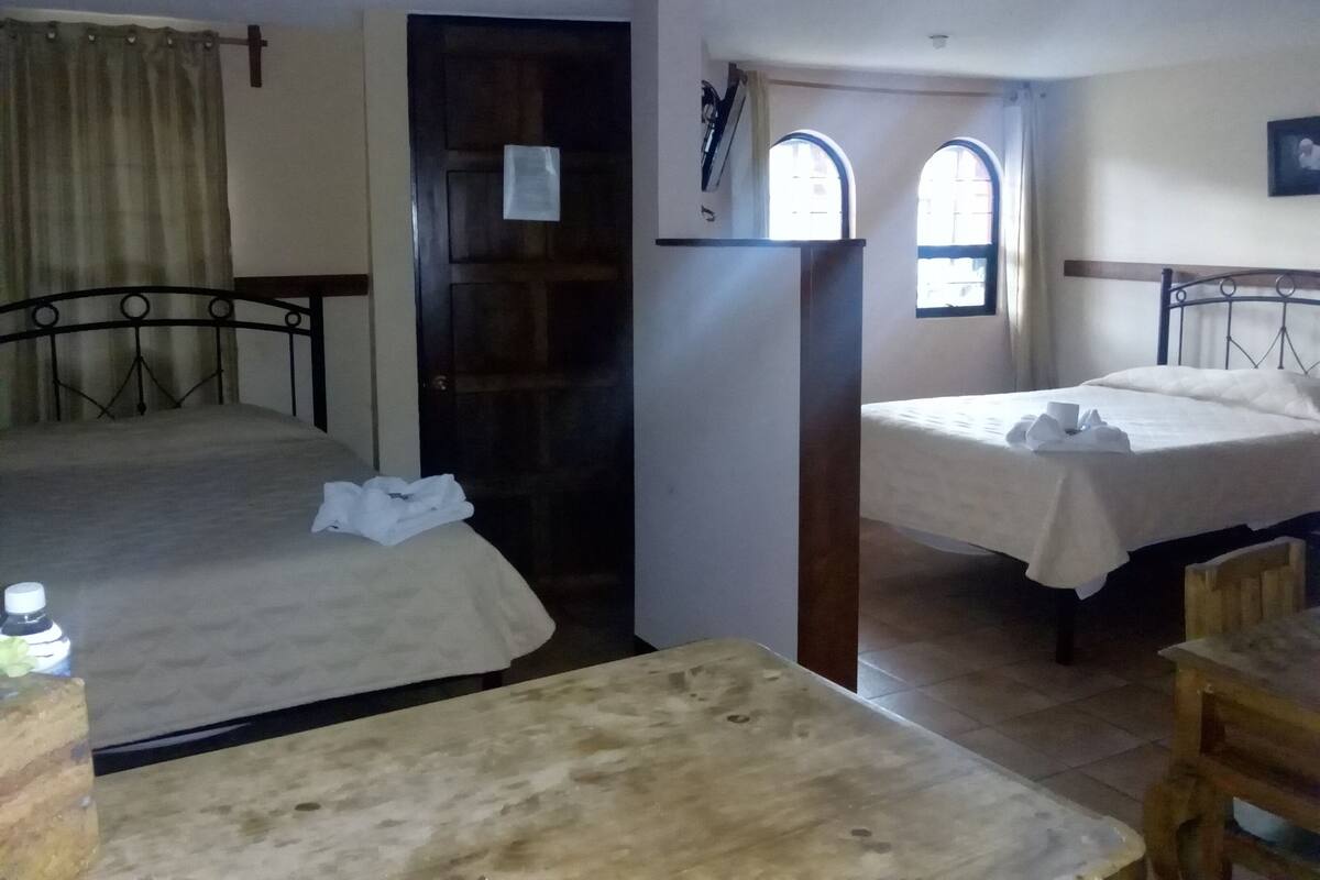 A small, dimly lit room with two single beds, a wooden table, and a door leading to another room.