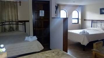 Una habitación pequeña y tenue con dos camas individuales, una mesa de madera y una puerta que da a otra habitación.