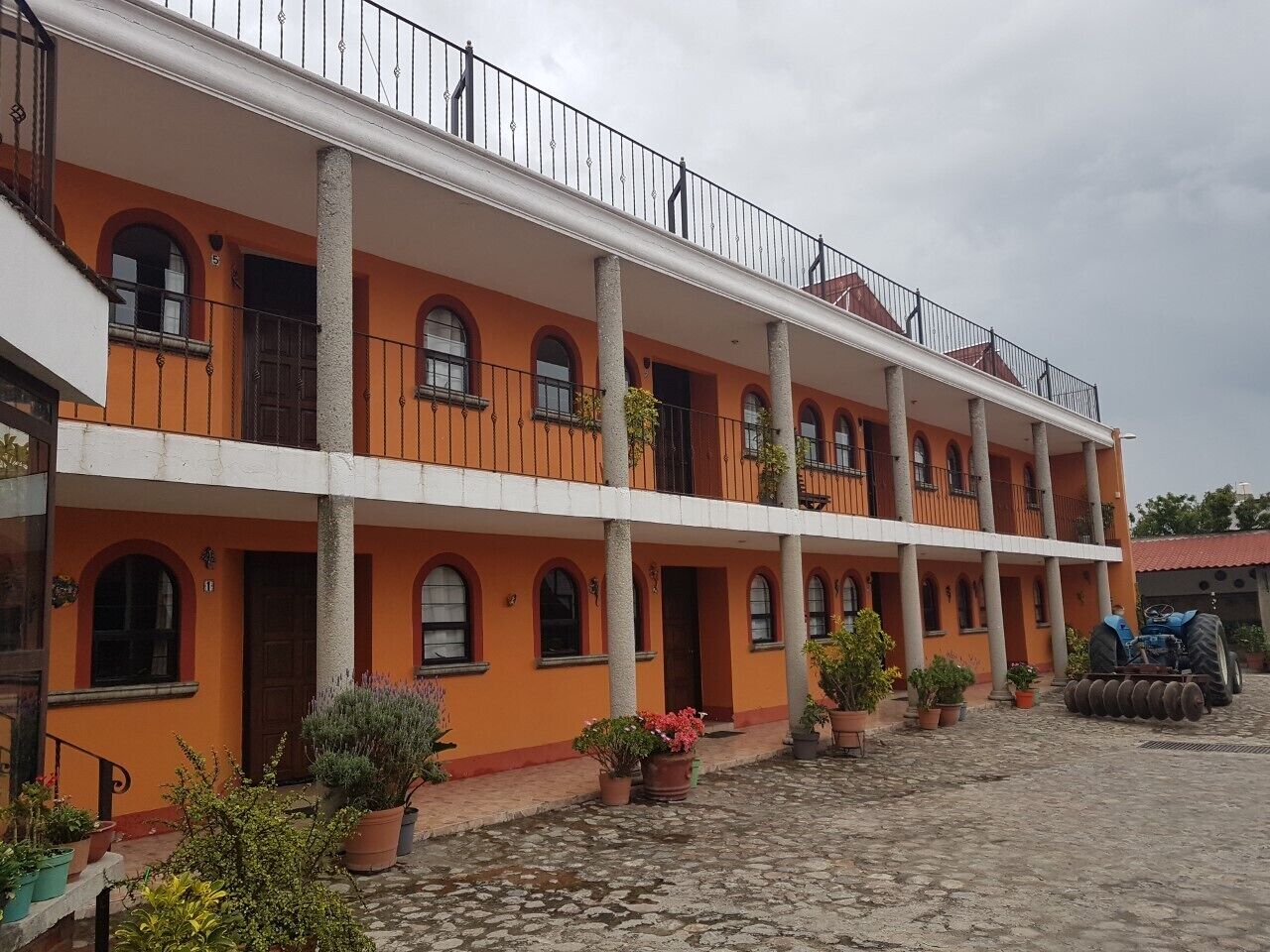Edificio de dos pisos con paredes naranjas y balcones blancos, que cuenta con un patio con plantas en macetas y un tractor estacionado.