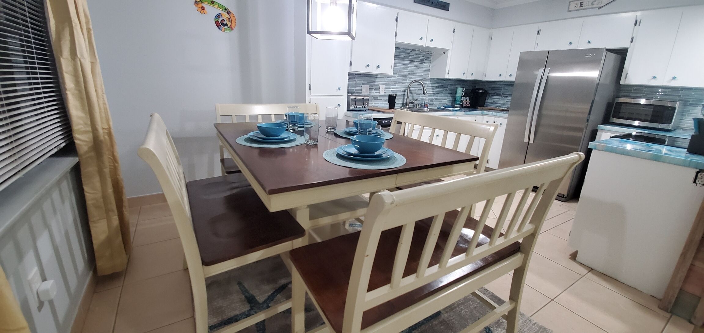 A dining area with a wooden table set for four, white chairs, and a kitchen in the background with white cabinets and stainless steel appliances.