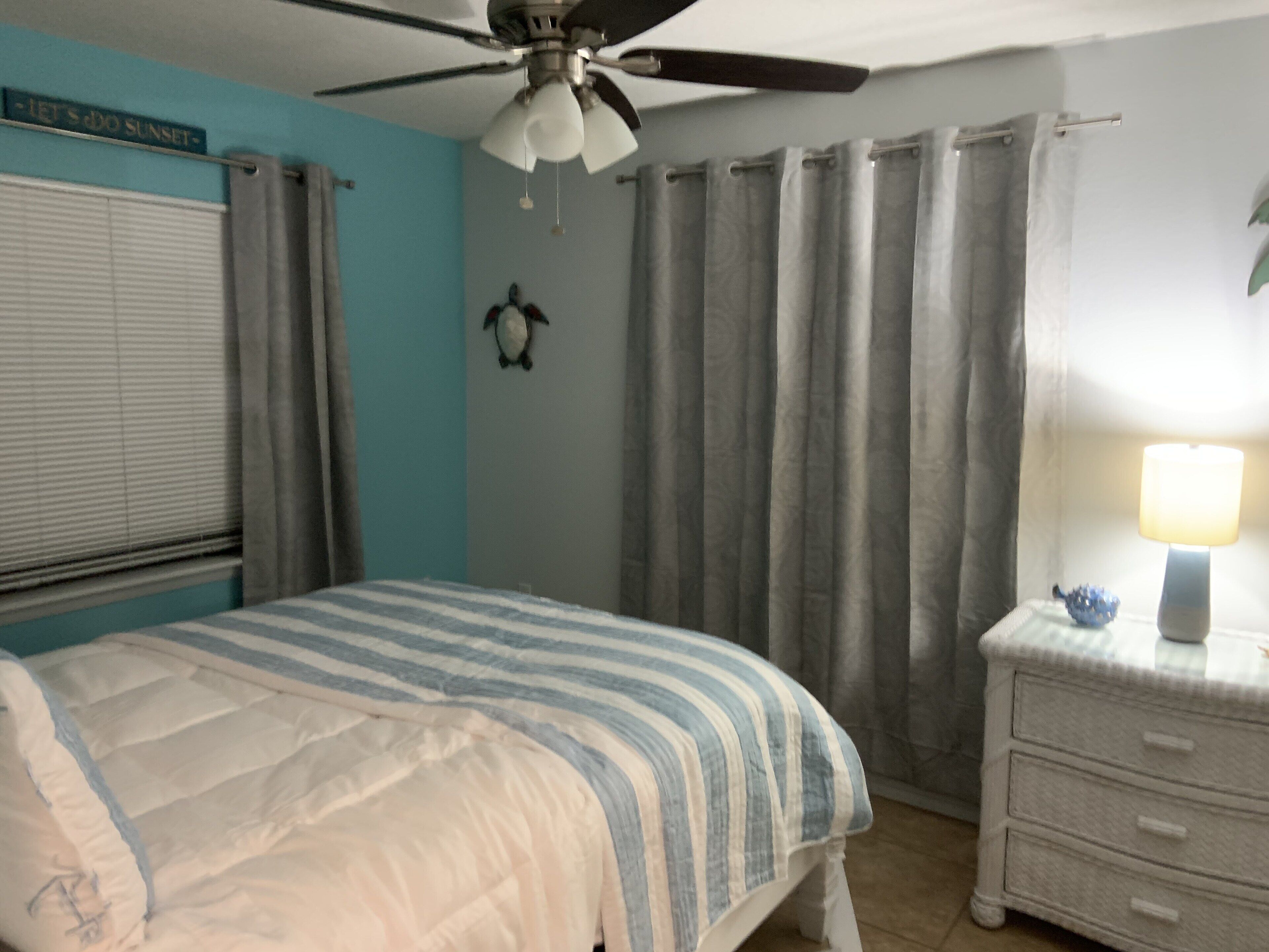 Un dormitorio con cama, mesita de noche, lámpara, ventilador de techo y cortinas.