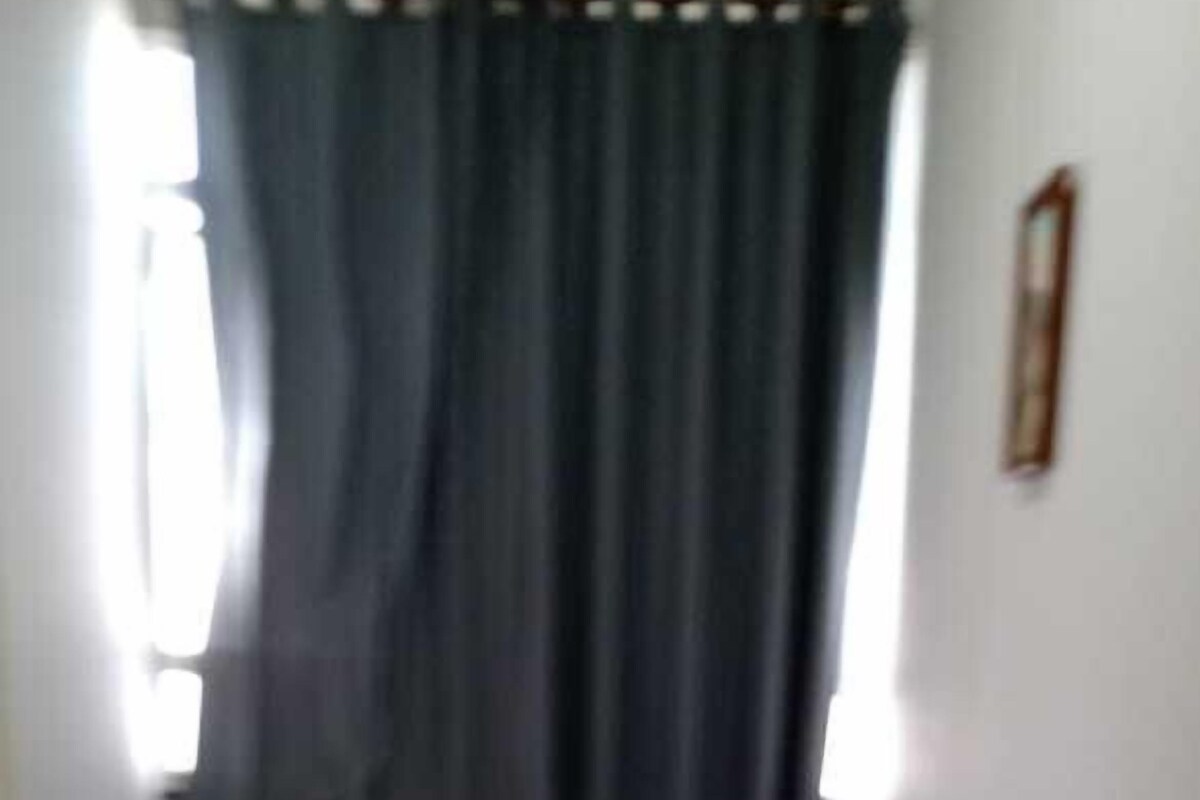 Una cama pequeña y bien hecha con sábanas y almohadas blancas, una ventana con cortinas oscuras y un radiador en la pared.