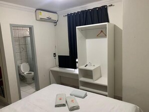 A hotel room with a bed, a desk with a computer, a wardrobe, a bathroom with a toilet, and an air conditioning unit.