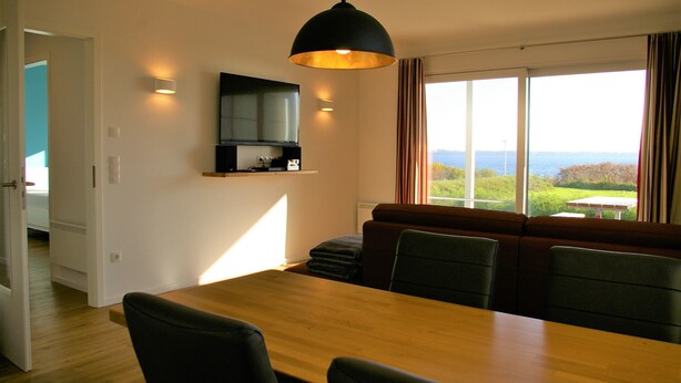 A dining area with a wooden table and chairs, a black pendant light, a built-in shelf with a television, and a large window with a view of greenery and a body of water.