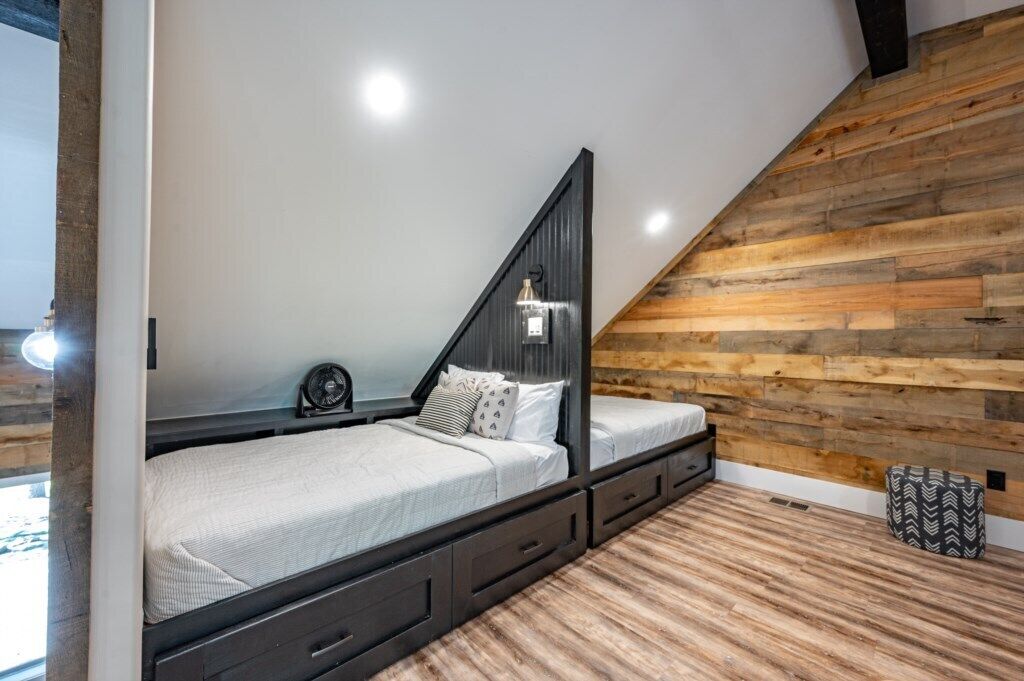 A modern loft bedroom with a sloped ceiling, exposed wooden beams, a built-in bed with storage drawers, and a small bedside table.