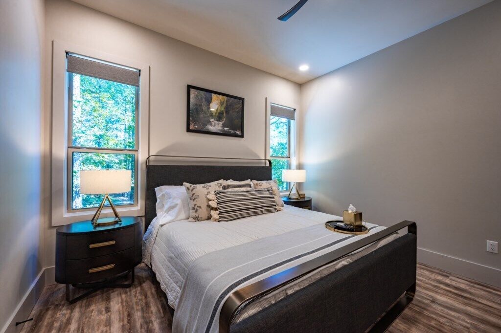 A bedroom with a large bed, two bedside tables, a nightstand, a window with a view of greenery, and a ceiling fan.