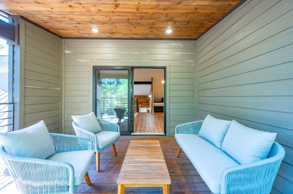 Un patio couvert, aménagé avec des meubles en osier et une table en bois, qui donne sur une porte vitrée menant à l’intérieur.
