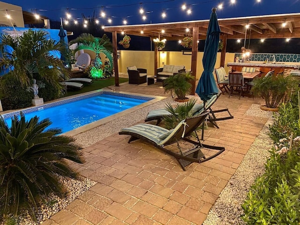 Een zwembadgedeelte met ligbedden, een pergola, sfeerverlichting en een bar.