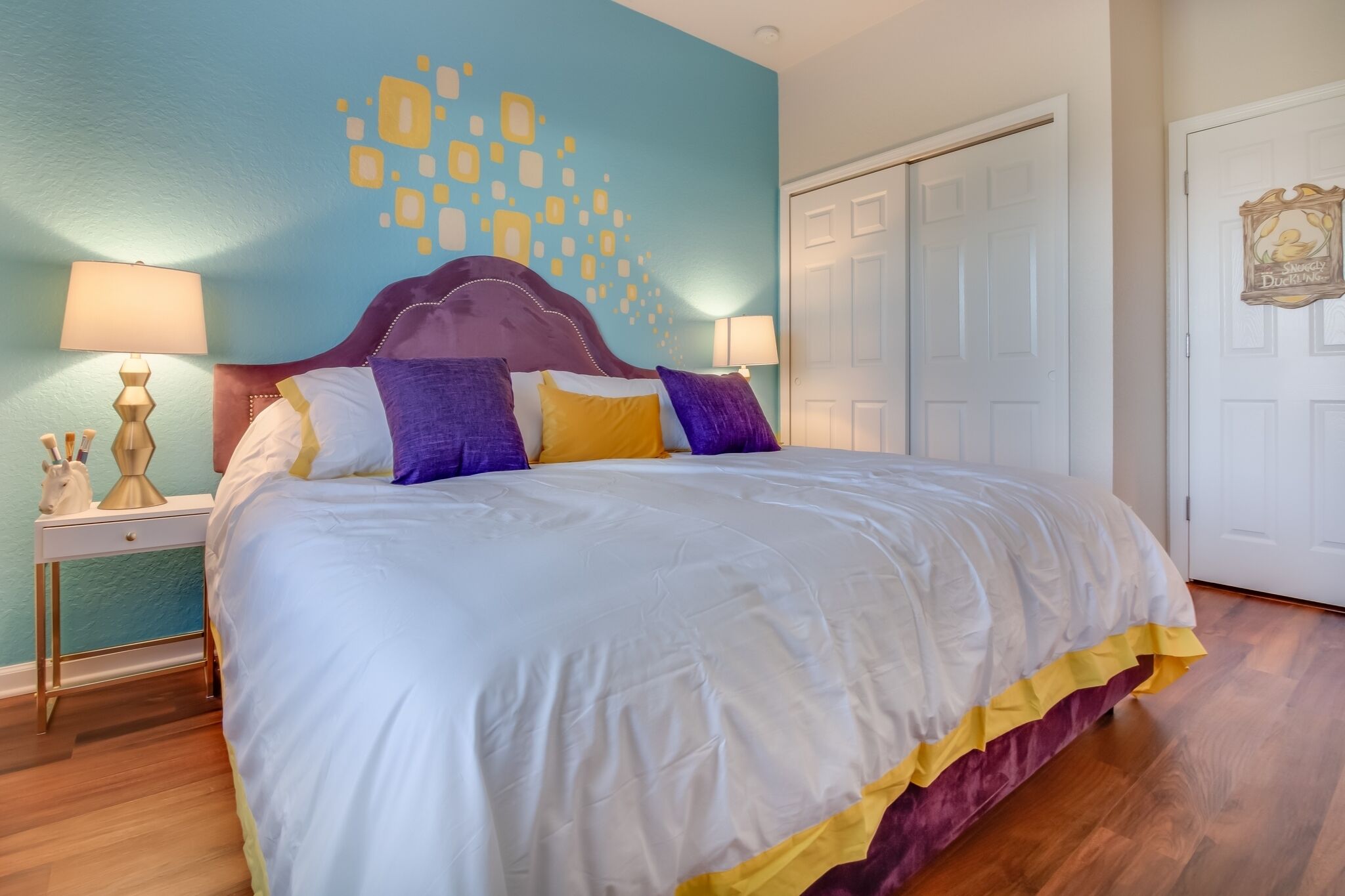 Un lit bien fait, recouvert d’une couvre-lit blanche et jaune, une table de chevet en bois avec une lampe, et un mur orné d’un motif géométrique.
