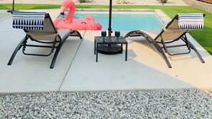 Área de piscina con dos sillones reclinables, una mesa con dos bebidas y una sombrilla grande que da sombra.