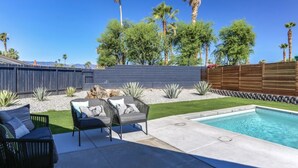 Un patio trasero con piscina, cerca de madera y zona verde con palmeras.