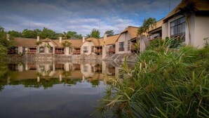 A row of bungalow-style buildings with thatched roofs, a calm water body in the foreground, and lush greenery around. - Umbhaba Lodge (Mbombela)