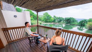 A wooden deck with a table and chairs overlooking a calm body of water and greenery. - Umbhaba Lodge (Mbombela)