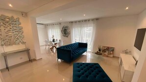 Una sala moderna con un sofá azul grande, un sillón azul más pequeño y una mesa de centro de vidrio. Se observa un mueble blanco y una pieza decorativa en la pared.