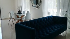 Una sala de estar moderna con un sofá azul oscuro, una mesa de centro de vidrio y una zona de comedor con sillas blancas y una mesa de madera.