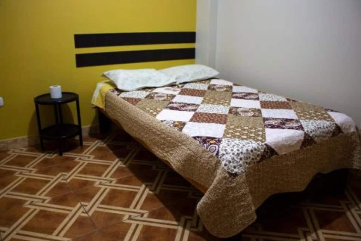 A single bed with a checkered quilt, a small bedside table with a roll of toilet paper, and a patterned floor.