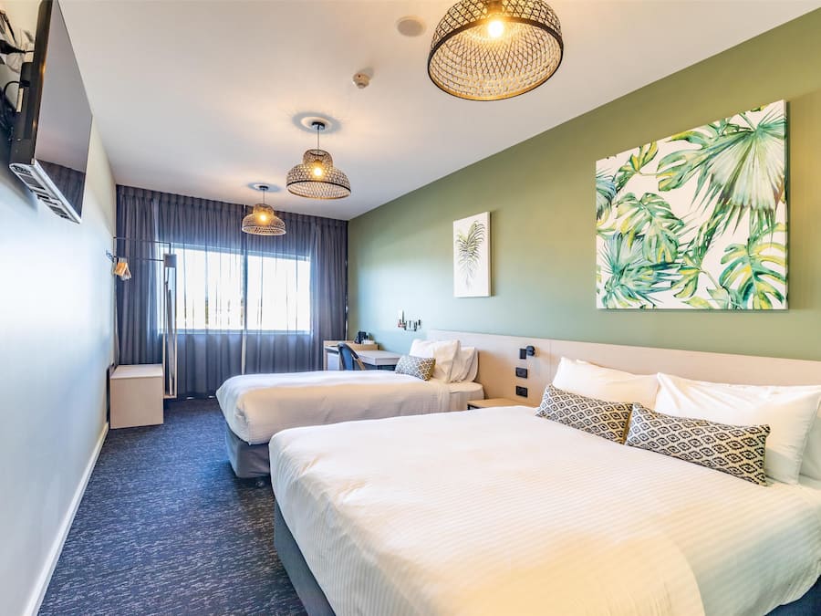 A hotel room with two beds, a desk, a chair, and a large tropical plant on the wall.