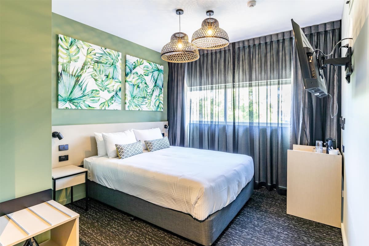 A hotel room with a bed, a nightstand, a desk, a wall-mounted TV, and a large window with curtains.