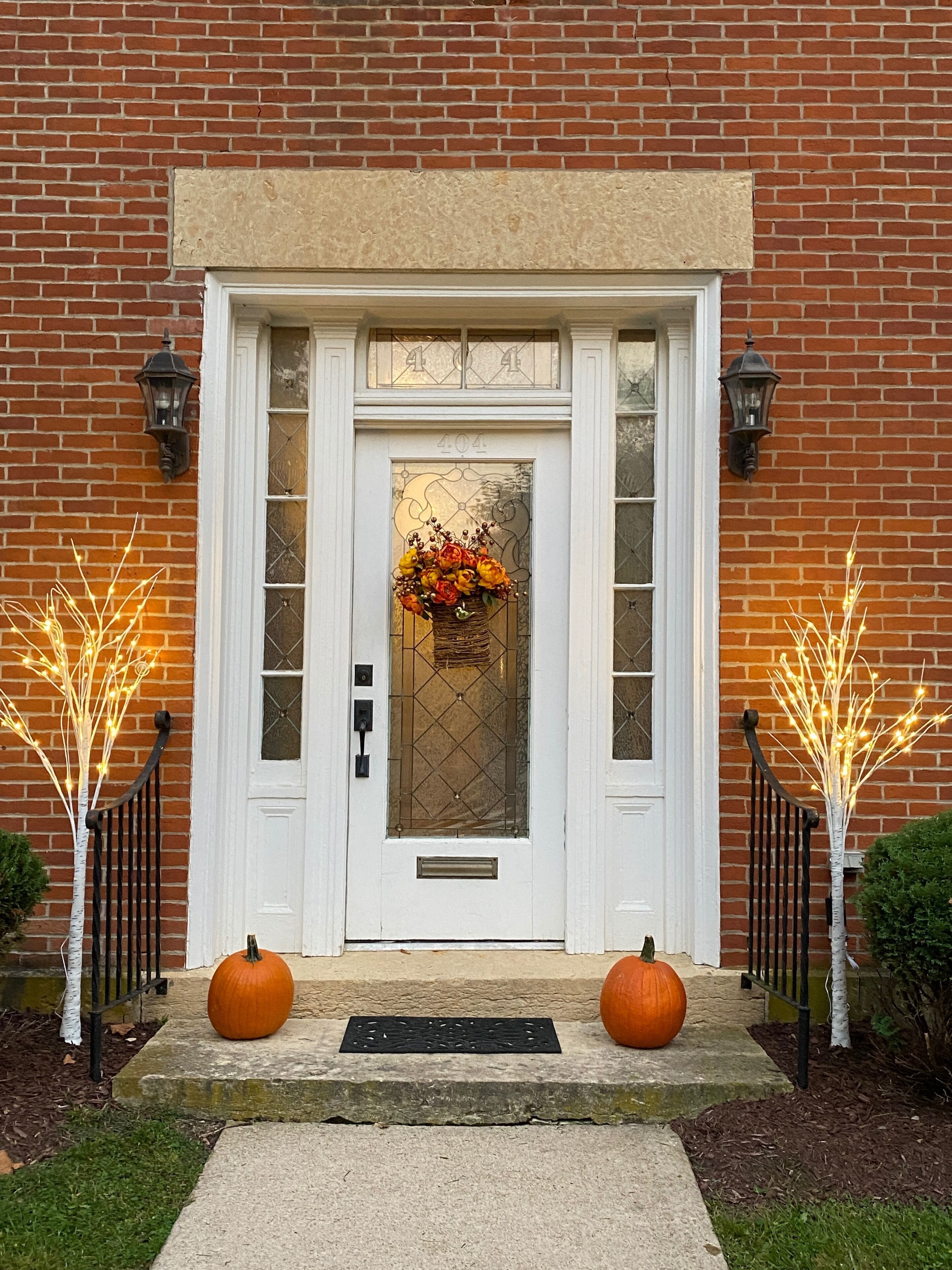 A front door with a number plate, a flower basket, and two pumpkins on the steps.