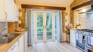 Una cocina con pared de piedra, vigas de madera, estufa a gas y vistas al jardín a través de un ventanal.