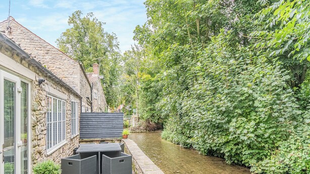 Un canal estrecho con un edificio de piedra en un lado y un seto verde exuberante en el otro. Hay un pequeño espacio al aire libre con dos sillas y una mesa.