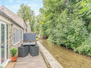 A narrow canal with a stone building on one side and a lush green hedge on the other. There is a small outdoor seating area with two chairs and a table.