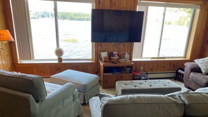 A cozy cabin-style room with a large window offering a view of a lake, a flat-screen TV mounted on the wall, and a wooden bench.