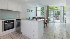 A modern kitchen with white cabinets, a built-in oven, and a dishwasher. There is a dining area with a table and chairs, and a view of the outdoors through large sliding glass doors.