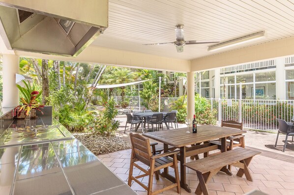 Une terrasse extérieure avec une table et des chaises en bois, entourée d’une végétation luxuriante et d’une clôture blanche.