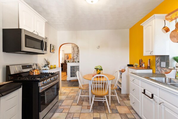 A compact kitchen with white cabinets, a stove, and a microwave. There's a dining area with a table and chairs, and a yellow accent wall. - Stunning 1 BR-Apt-Oceanview to Revere Beach -3 Train Stop Boston Logan Airport (Revere)