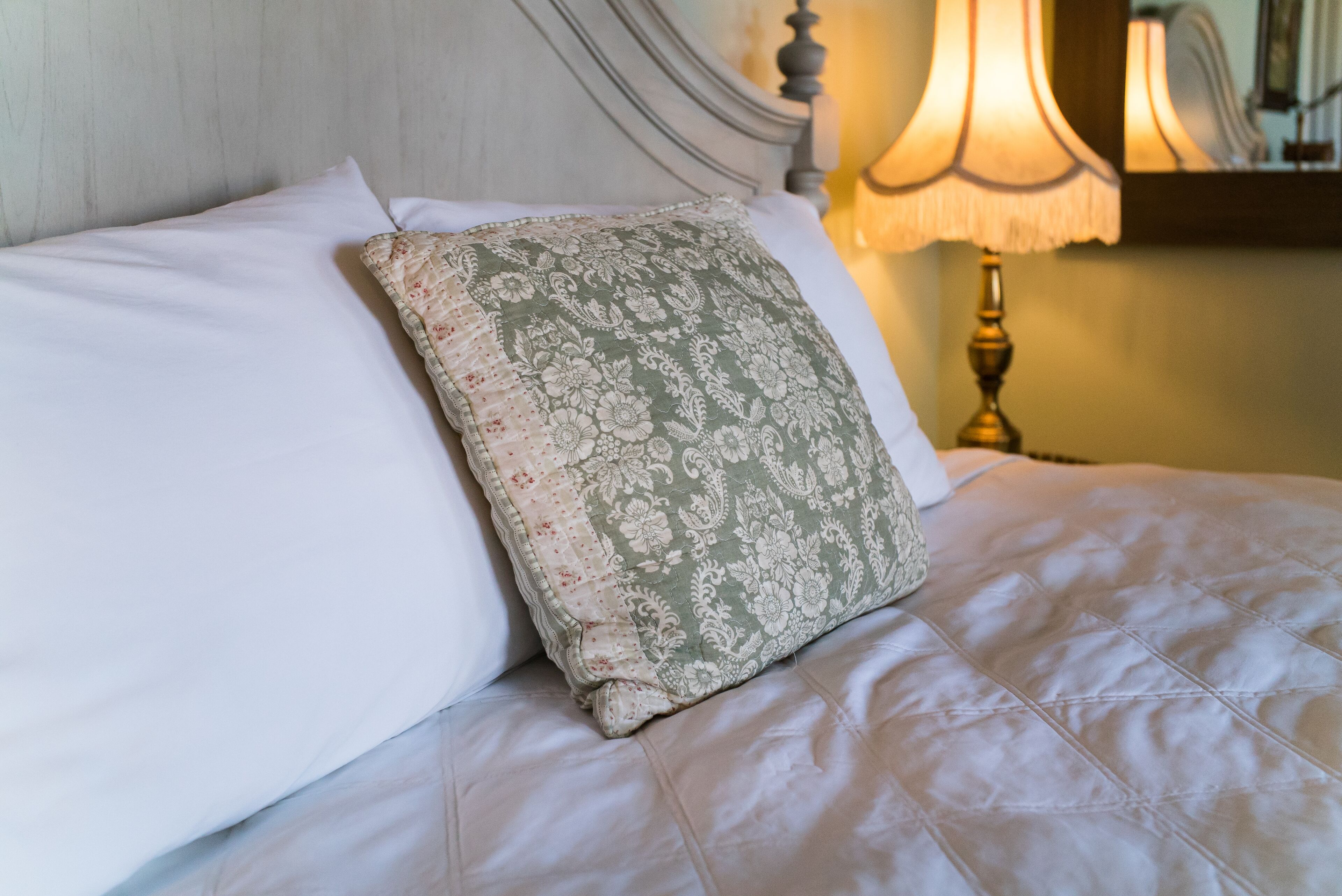 A neatly made bed with a floral pillow and a white pillow, a bedside lamp, and a wooden headboard.