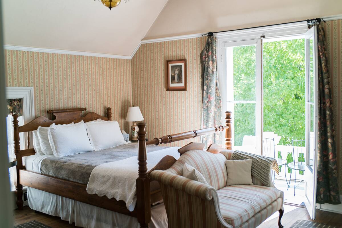 A bedroom with a large bed, a wooden headboard, a bedside table with a lamp, and a view of the outdoors through a window.