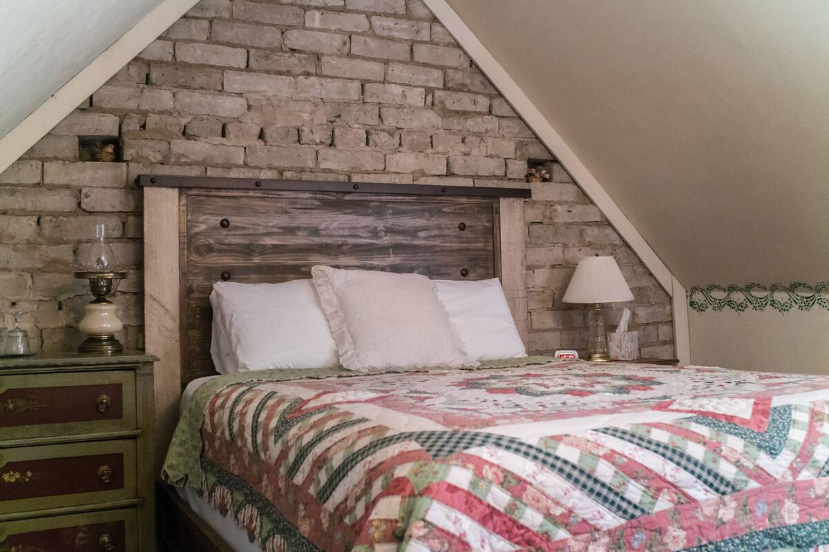 A bedroom with a brick wall, a wooden headboard, a quilted bedspread, a nightstand with a lamp, and a dresser with drawers.