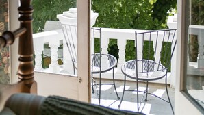 Uitzicht door een raam op een balkon met een witte railing, een metalen stoel en een witte tafel.
