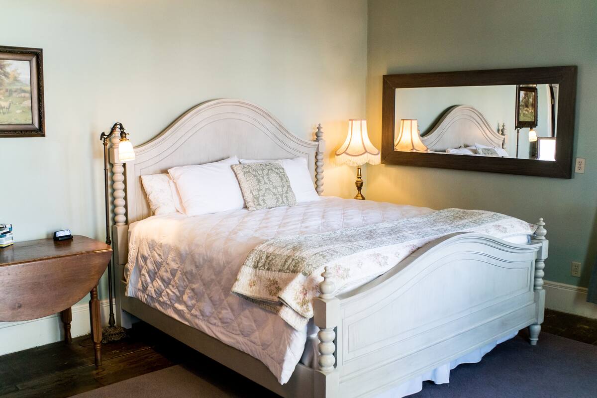 A neatly made bed with a quilt, a bedside lamp, and a nightstand with a lamp. A large mirror hangs above the bed.