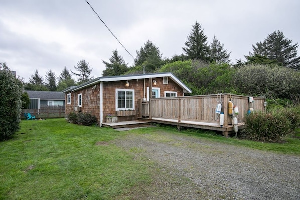 A wooden cabin with a deck, surrounded by a grassy yard and dense trees. - Sand Bucket Cottage (Pacific Beach)
