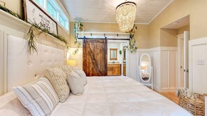 Une chambre à coucher avec un grand lit, un lustre, une porte en bois et une salle de bain visible à travers une porte ouverte.