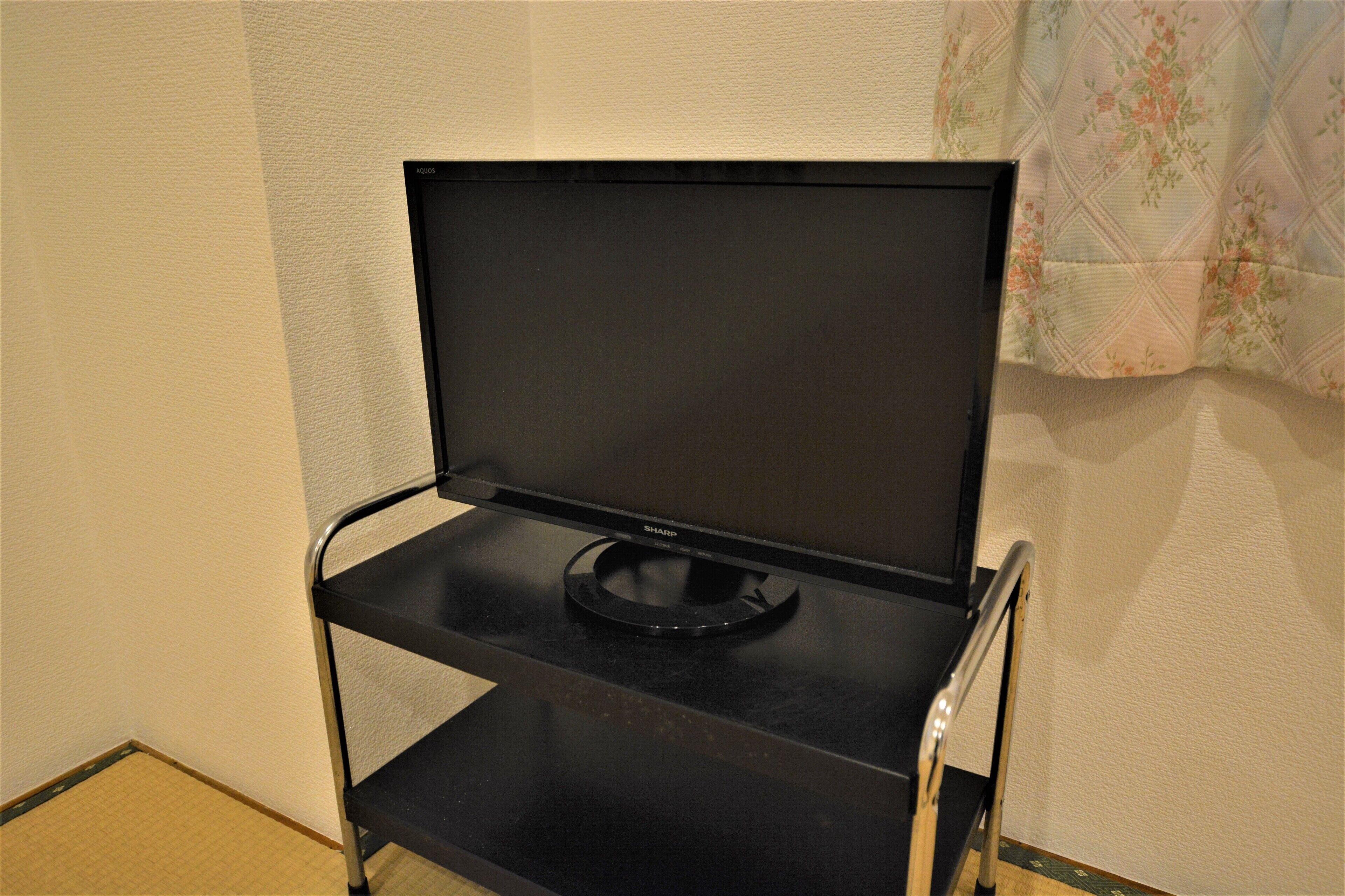 A black TV on a metal stand in a room with a plain wall and a floral curtain.