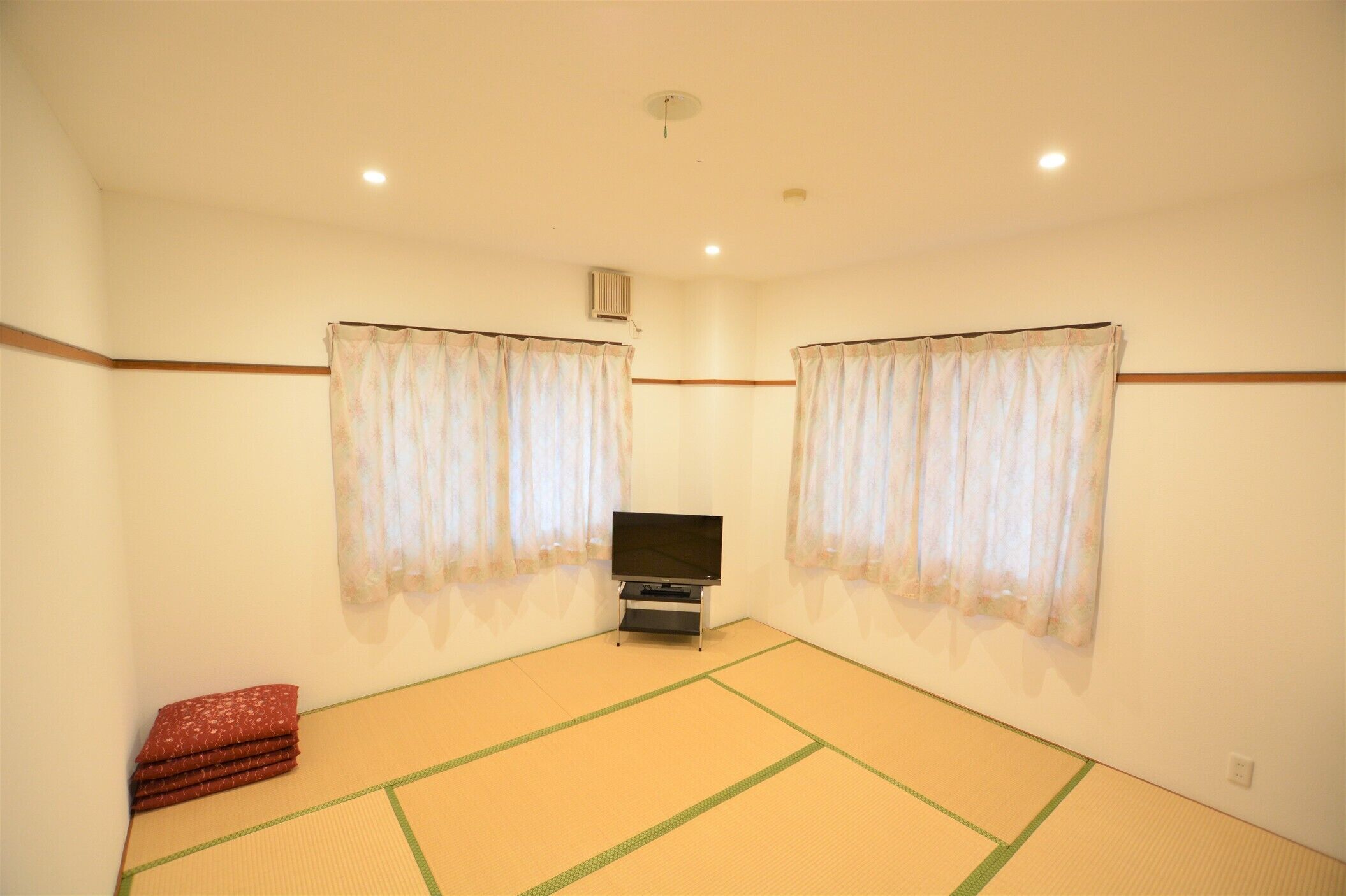 A room with tatami mat flooring, a television, and two windows with curtains.