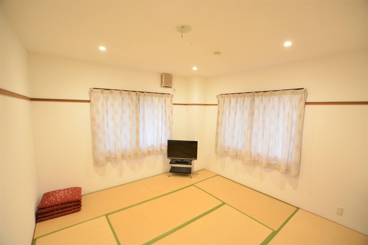 A room with tatami flooring, a television, and two windows with curtains.