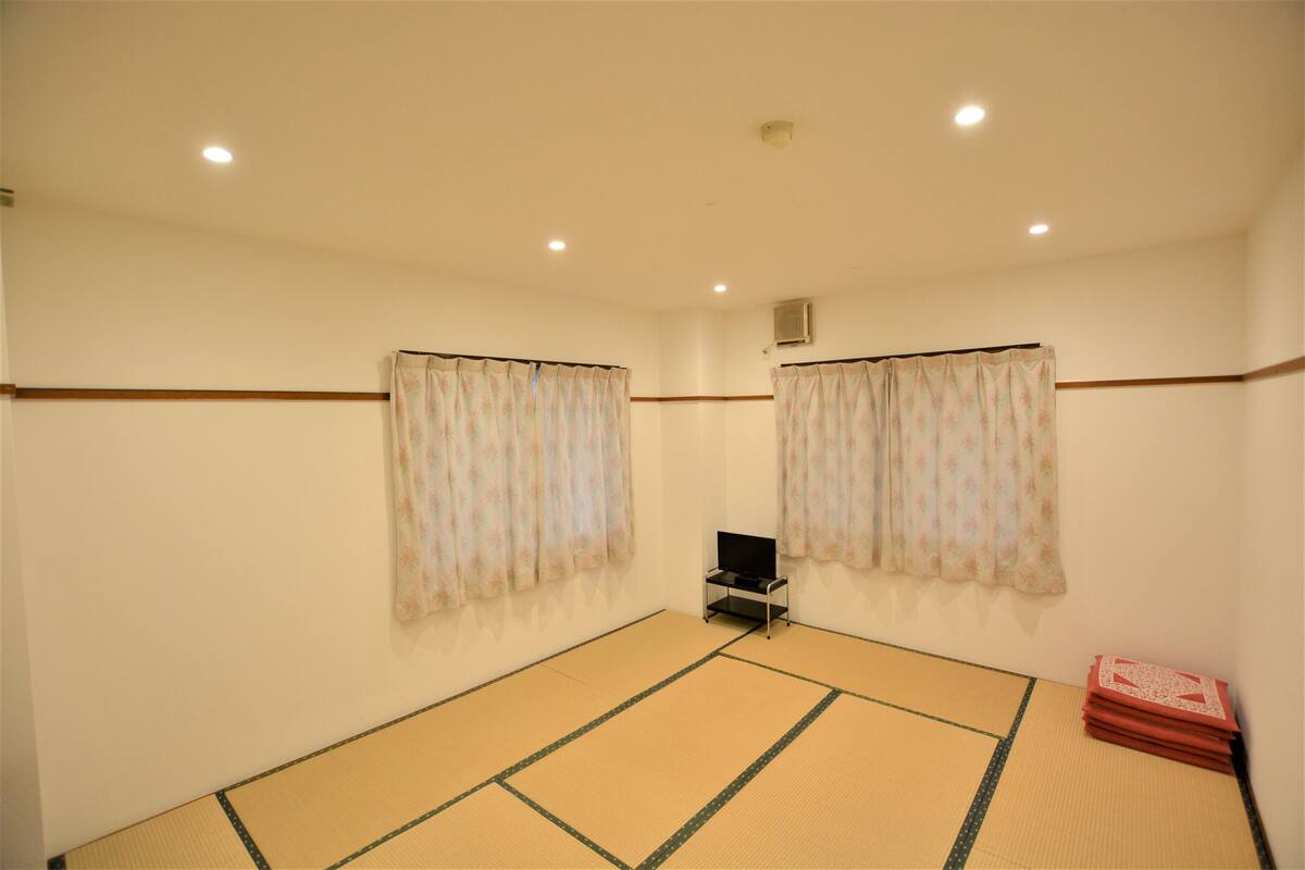 A room with tatami flooring, a television on a stand, and a stack of books.