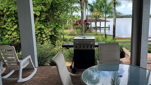 Un espace patio avec une table en verre, deux chaises, un barbecue et une vue sur un lac, avec un quai et des palmiers.