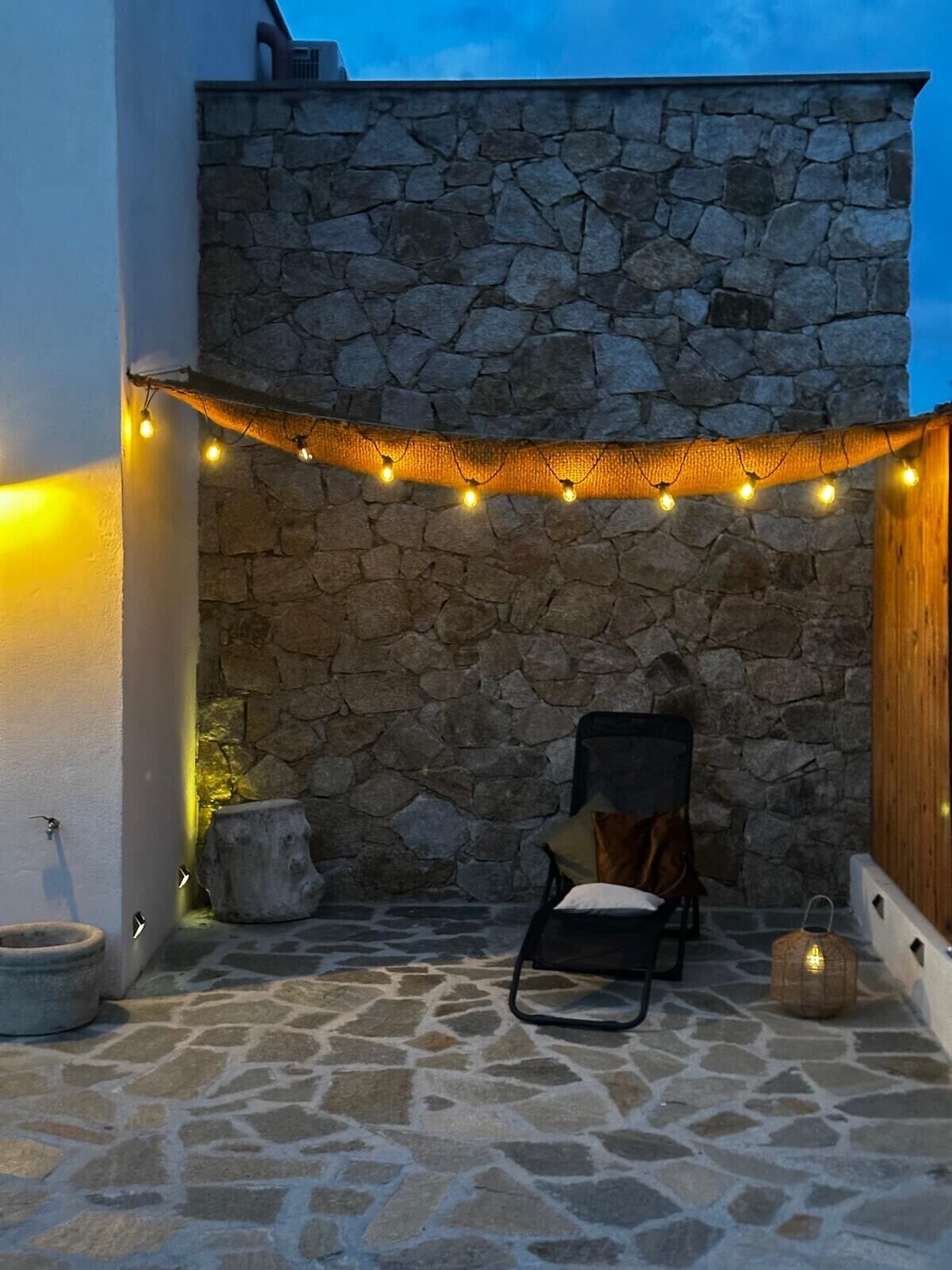 Un mur de pierre orné d’une guirlande lumineuse, une chaise et une petite table dans une cour.