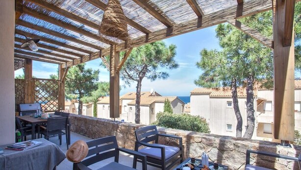 Un patio al aire libre con mobiliario de madera, una mesa con un sombrero y vistas a edificios y árboles.