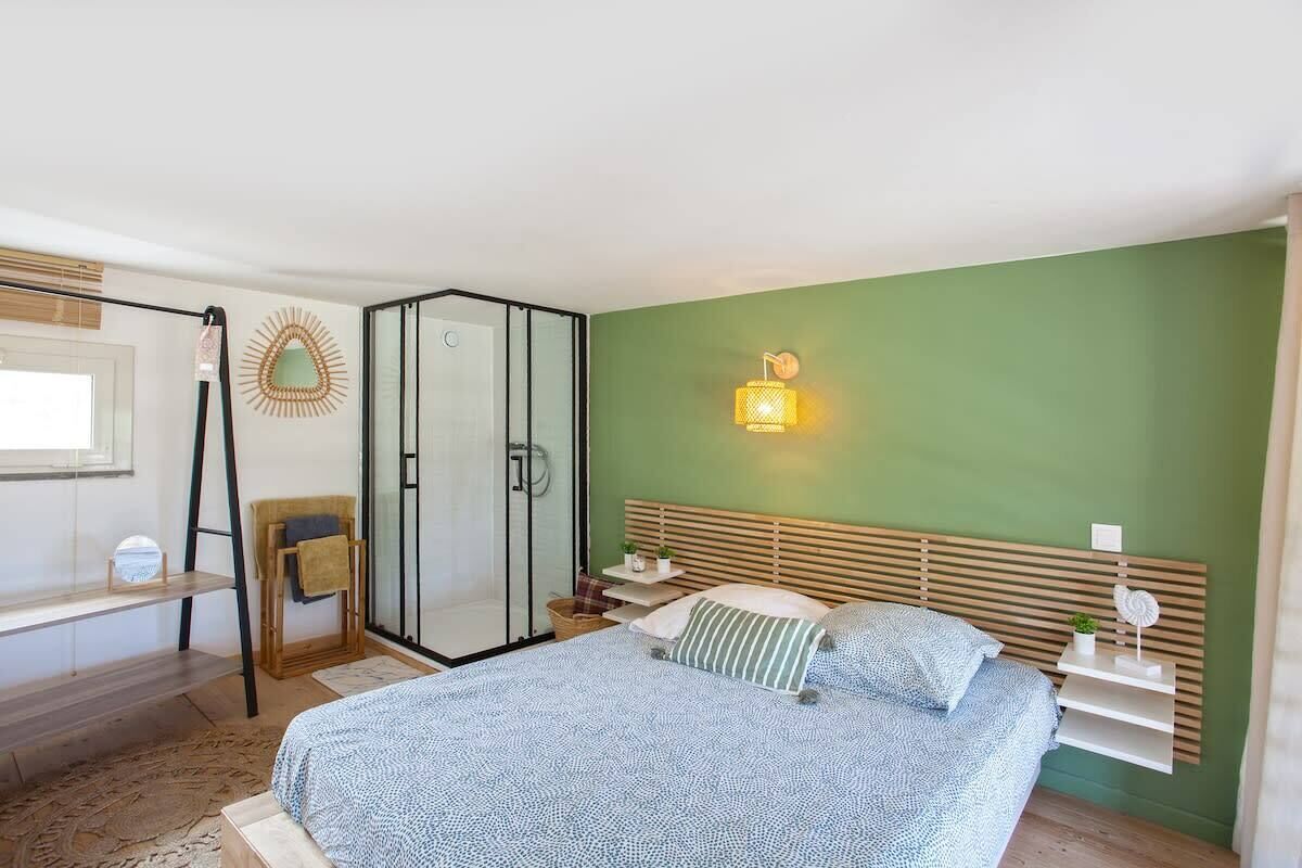 A bedroom with a bed, a nightstand, a bedside table, a window, a mirror, a towel rack, and a shower area.