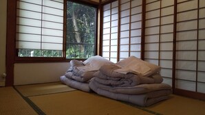 A tatami mat room with a large window, a stack of bedding, and shoji screens. - Hakone Guesthouse toi - Hostel (Hakone)