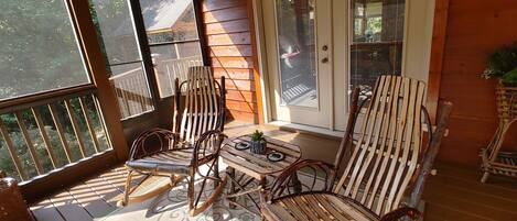 A wooden porch with two rocking chairs and a small table, a door with a 'TIGHT' sign, and a view of greenery outside.